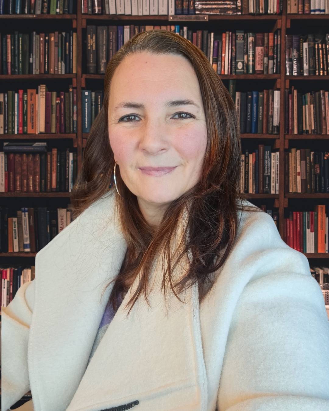 Portrait of Victoria, creator of Letters from Mars, smiling in a light-colored coat in front of a large bookshelf filled with vintage books.