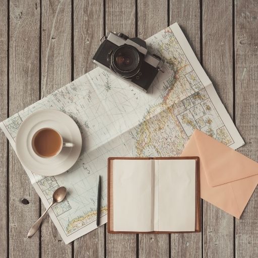 Top-down view of travel and writing artifacts on a rustic wooden table, featuring an open paper map, a vintage film camera, a cup of coffee, a blank notebook with a pen, and a peach-colored envelope.