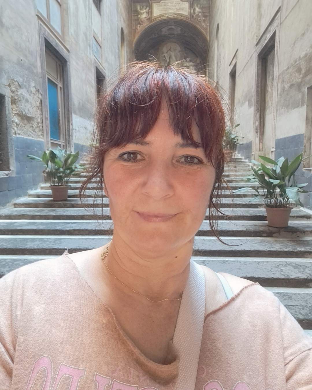 lose-up selfie portrait of Victoria, the creator of Letters from Mars, smiling. She is standing at the bottom of a wide stone staircase within a historic European building corridor with potted plants.