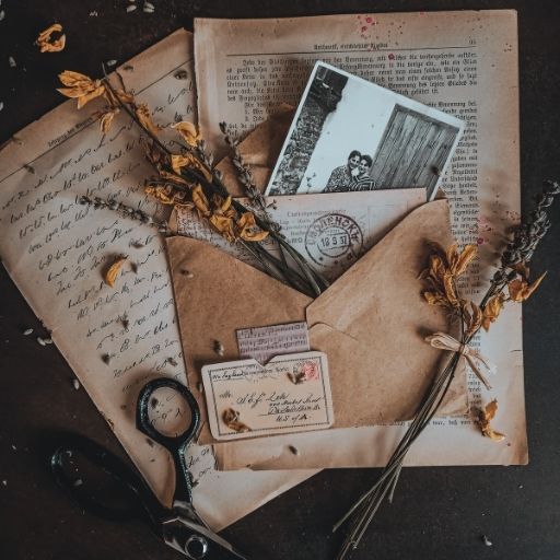 A top-down still life of old handwritten documents, a kraft paper envelope containing a black and white photograph of a couple, dried lavender, and vintage scissors on a dark tabletop.